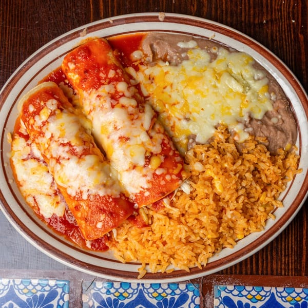 Happy Mexican enchiladas with red sauce, melted cheese, rice, and refried beans on a plate.